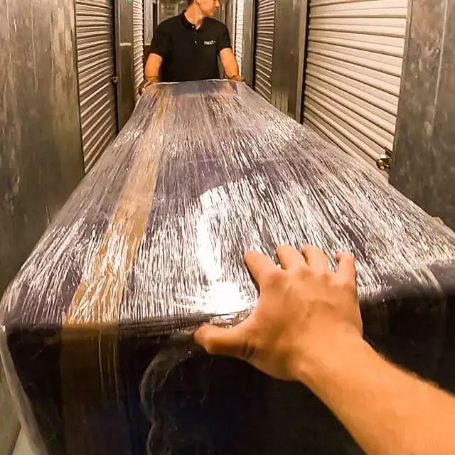 Two Next Moving local movers roll a couch protected with moving blankets and shrink wrap inside storage facility among storage units near me.